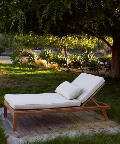 The Outdoor Vista Lounge Chair, featuring a cushion and pillow, is placed on a wooden deck in a shaded garden filled with greenery and sunlit plants beneath a large tree.