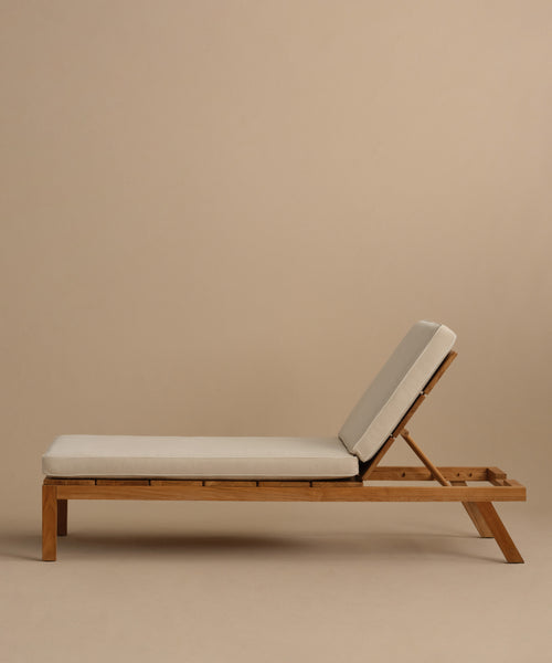 The Outdoor Vista Lounge Chair features a minimalist teak frame and light beige cushions, set against a neutral beige background. Its angled backrest offers comfortable reclining, making it an inviting and durable outdoor furniture piece.