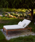 The Outdoor Vista Lounge Chair with white cushions rests on a wooden platform in a grassy garden, shaded by trees and surrounded by plants and sunlight—a perfect, durable spot for outdoor relaxation.