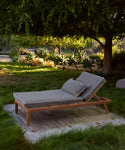 The Outdoor Vista Lounge Chair with a plush pillow sits on a wooden platform in a grassy yard, shaded by a large tree and surrounded by vibrant plants.