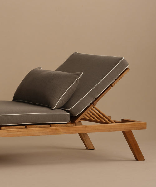 The Outdoor Vista Lounge Chair features a modern teak frame with angled legs, a gray cushioned seat, matching pillow, and white piping, all set against a neutral beige background.