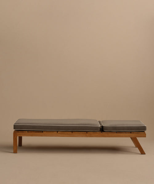 The Outdoor Vista Lounge Chair is a minimalist wooden daybed with two gray cushions, set against a plain beige background. Its simple, modern form and slightly elevated side are inspired by the timeless outdoor lounge chair design.