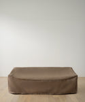 A large, rectangular object protected by the Outdoor Vista Sofa Cover in smooth brown rests on a light wooden floor against a plain white wall.