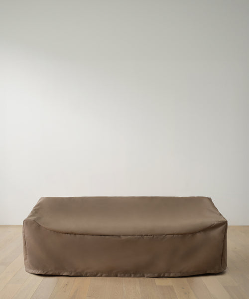A large, rectangular object protected by the Outdoor Vista Sofa Cover in smooth brown rests on a light wooden floor against a plain white wall.