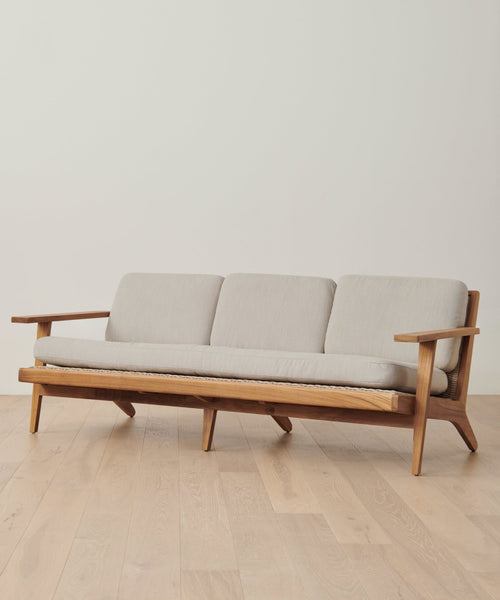 A modern wooden sofa with light beige cushions and three seats on a light wood floor against a white wall—perfectly protected by the Outdoor Vista Sofa Cover for stylish patio furniture care.