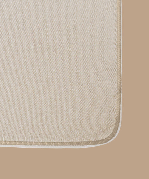Close-up of the corner of an Outdoor Vista Sofa Cushion in beige, with a slightly darker beige border, set on a solid light brown surface.
