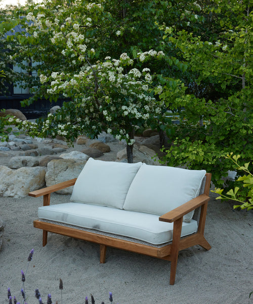 The Outdoor Vista Sofa with Tempotest fabric cushions sits on sandy ground in a garden, surrounded by rocks, green trees, and blooming white flowers.