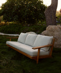 The Outdoor Vista Sofa, featuring Tempotest fabric cushions, sits on green grass near a large rock, surrounded by trees and bushes in the background.