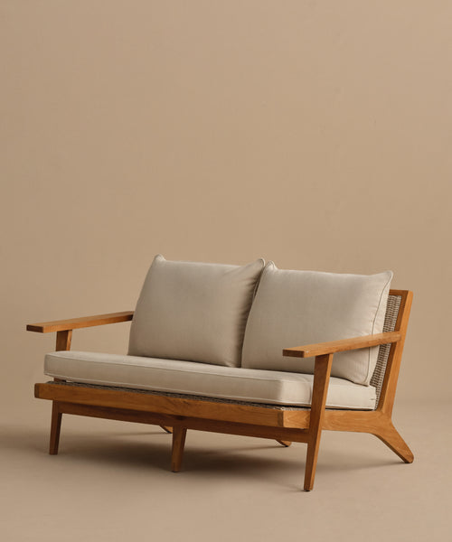 The Outdoor Vista Sofa features angled armrests and light beige Tempotest fabric cushions, crafted in modern teak and displayed against a plain beige background.