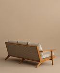 The Outdoor Vista Sofa, featuring a wooden frame, woven backrest, and beige Tempotest fabric cushions, is shown angled against a plain tan background. Its minimalist style reflects mid-century modern design.
