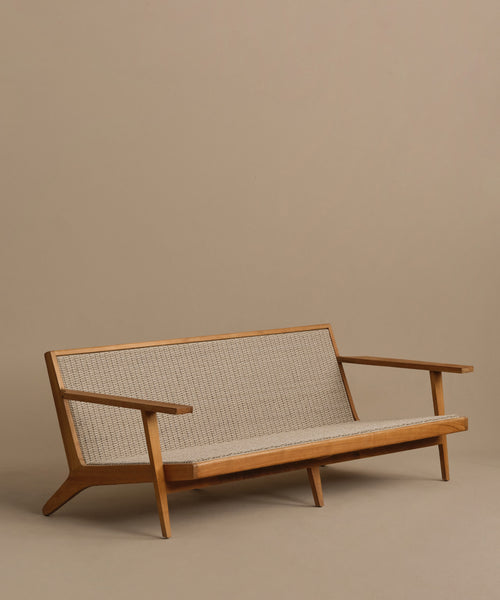 The Outdoor Vista Sofa features a minimalist design with angled legs, wide armrests, and a woven beige back and seat, shown against a plain light brown background.