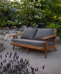The Outdoor Vista Sofa with gray cushions is placed on sandy ground in a garden, surrounded by blooming flowers, leafy green trees, and large rocks.