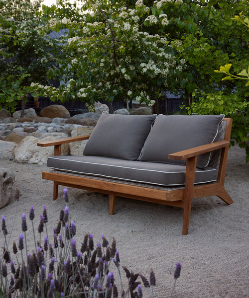 The Outdoor Vista Sofa with gray cushions is placed on sandy ground in a garden, surrounded by blooming flowers, leafy green trees, and large rocks.