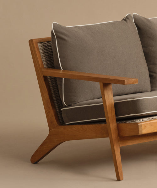 A close-up of the Outdoor Vista Sofa, a modern teak armchair with gray cushions and white piping, set against a plain beige background.