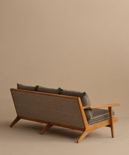 The Outdoor Vista Sofa, a mid-century modern teak piece with angled legs, dark gray cushions, and a textured backrest, is shown from behind against a plain beige background—ideal for stylish outdoor spaces.