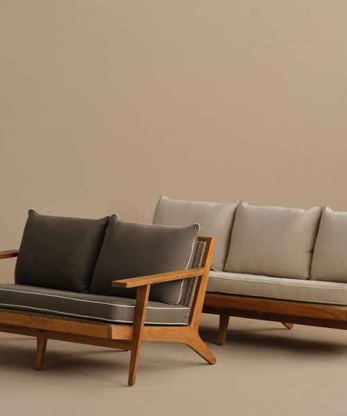 Two Outdoor Vista Sofas with angled legs—one with dark gray cushions in front and another with light beige cushions behind—are set against a plain beige backdrop.