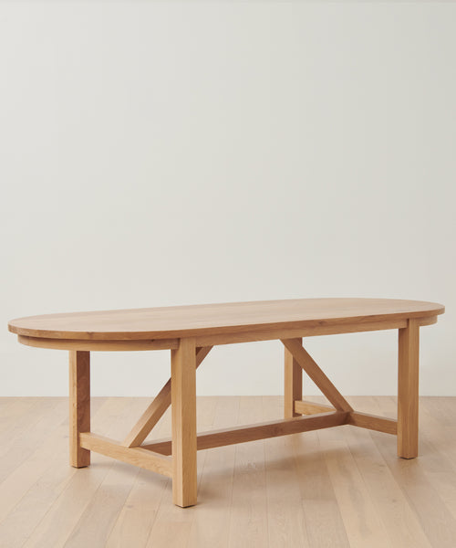 The Montecito Oval Dining Table, handmade from solid white oak with sturdy legs and a crossbar support, sits elegantly on a light wood floor against a plain white wall.