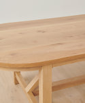 A close-up of the Montecito Oval Dining Table shows its solid white oak build, rounded edges, visible wood grain, and thick legs. The handmade table features a simple, sturdy design and sits atop a matching wooden floor.