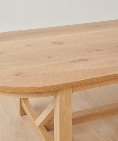 A close-up of the Montecito Oval Dining Table shows its solid white oak build, rounded edges, visible wood grain, and thick legs. The handmade table features a simple, sturdy design and sits atop a matching wooden floor.
