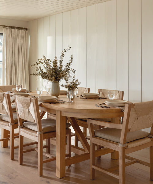 The Montecito Oval Dining Table, crafted from solid white oak, is paired with six cushioned chairs and set with plates, glasses, and utensils. A vase of dried flowers sits at the center amid light walls, wood floors, and natural curtains.
