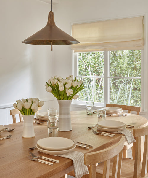 A bright dining room features the Montecito Oval Dining Table set for four with beige plates, glassware, striped napkins, and vases of white tulips by a window with a cream Roman shade and a modern pendant light overhead.