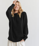 A blonde woman smiles with her eyes closed, wearing the Oversized Cotton Fisherman sweater in black and white pants. One hand is in her pocket, the other touches her head. She stands against a plain, light-colored background.