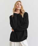 A woman with long blonde hair wears the Oversized Cotton Fisherman sweater in black and white pants, standing against a plain light background. She faces the camera with a relaxed expression, her left hand gently touching her shoulder.