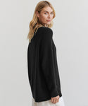 A woman with long blonde hair models the Oversized Cotton Fisherman sweater in black, posing slightly turned to the side against a plain light background.