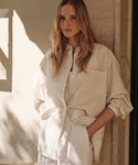 A woman with long, wavy blonde hair stands against a stone wall in soft sunlight, wearing the Oversized Shirt in cream paired with white pants.