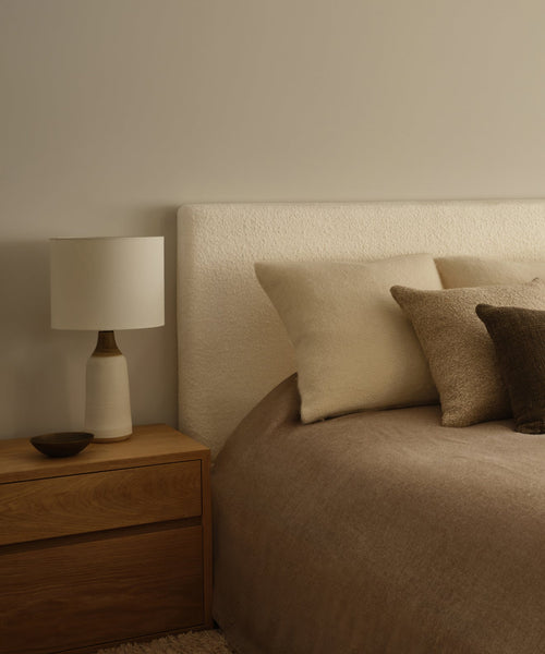 A minimalist bedroom featuring the Pacific Bed with an upholstered headboard, a wooden nightstand, a white table lamp, a small bowl, beige and brown pillows, and a taupe blanket.