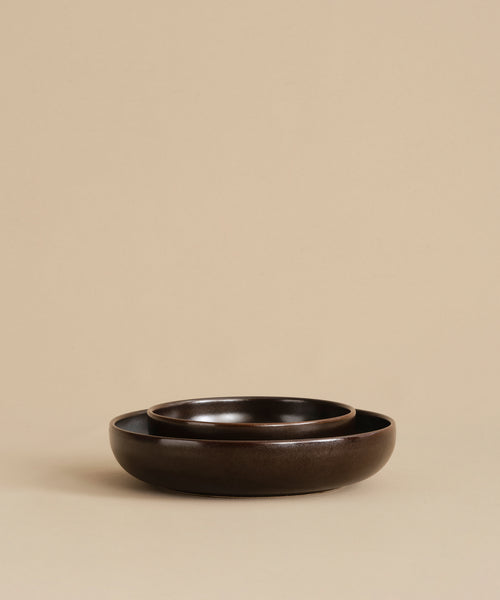 Two Pacific Breakfast Bowls in dark brown stoneware are nested together on a beige background, showcasing their minimalistic, elegant design and rich chocolate tones.