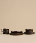 A set of dark brown ceramic dishes, featuring a Pacific Breakfast Bowl, two Dark Chocolate mugs, extra bowls, and plates, is neatly arranged on a beige surface against a beige background.
