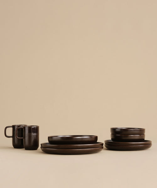 A set of dark brown ceramic dishes, featuring a Pacific Breakfast Bowl, two Dark Chocolate mugs, extra bowls, and plates, is neatly arranged on a beige surface against a beige background.
