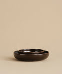 Two dark brown Pacific Soup Bowls, with a smaller one nested inside a larger, rest on a beige surface and matching background. Their minimalist arrangement highlights the elegant design of this dinnerware set.