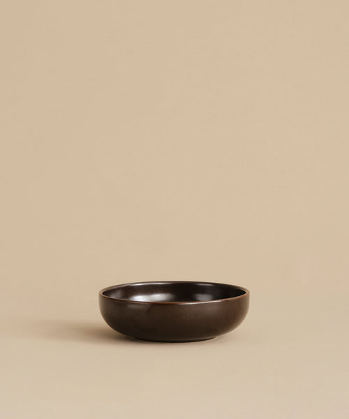 A small, round, dark brown Pacific Breakfast Bowl sits empty and centered on a beige surface with a plain beige background, highlighting its elegant simplicity.