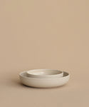 Two minimalist, round Pacific Breakfast Bowls in off-white—one nested inside the other—rest elegantly on a beige surface, set against a beige background. Modern dinnerware for any setting.