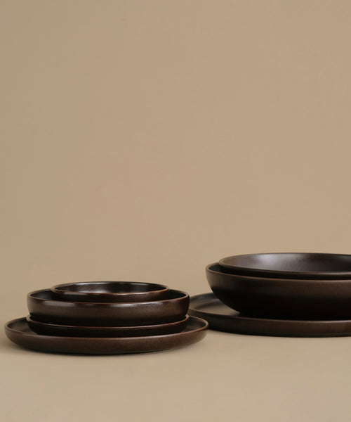 A set of dark brown ceramic plates and bowls with a matte, rustic finish, arranged on a solid beige background.
