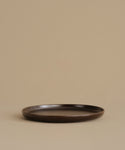 A dark brown ceramic dinner plate with a matte finish, photographed against a solid beige background.