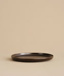 The Pacific Dinner Plate, a round, shallow, dark brown stoneware plate, is centered on a beige surface and background. Its elegant design makes it a perfect addition to any dinnerware collection. The plate is empty and tastefully displayed.