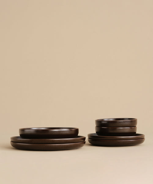 A set of dark brown ceramic dinnerware, including plates and a Pacific Soup Bowl, is neatly stacked on a light beige surface against a matching beige background.