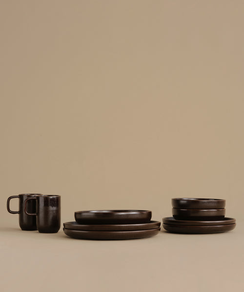Dark brown ceramic tableware set including plates, bowls, and mugs, arranged on a plain beige studio background.