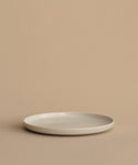 The Pacific Dinner Plate, a simple round white stoneware piece, rests on a beige surface against a matching beige background for a minimalist and neutral look.