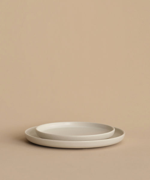 Two simple, round Pacific Dinner Plates in white stoneware are stacked against a beige background. Their minimalist design and soft lighting create a clean, modern look for any setting. Made in Portugal.