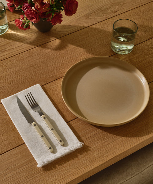 The Pacific Dinner Plate rests on a wooden table with a glass of water, white napkin, and cutlery. A bouquet of red flowers in the background completes this stylish Pacific Dinner Plate scene.