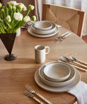 A round wooden table set with Pacific Dinnerware, featuring a Pacific Mug, cream-colored dishes and bowls. White-handled cutlery rests on napkins, and a vase of white tulips decorates the table as sunlight streams through sheer curtains.