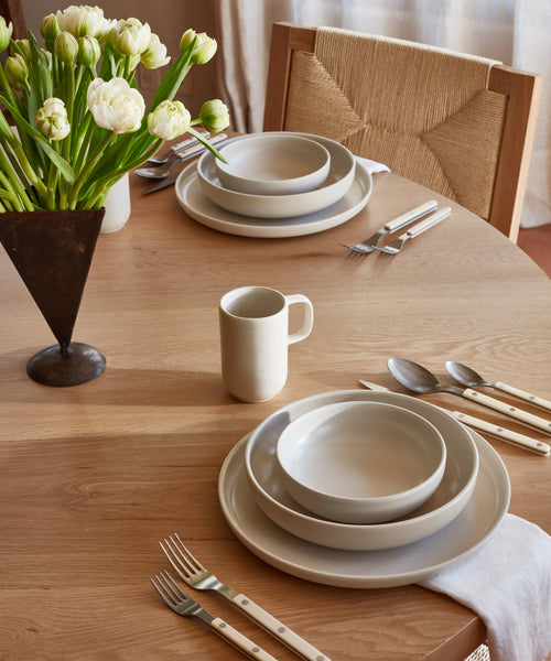 A round wooden table set with Pacific Dinnerware, featuring a Pacific Mug, cream-colored dishes and bowls. White-handled cutlery rests on napkins, and a vase of white tulips decorates the table as sunlight streams through sheer curtains.