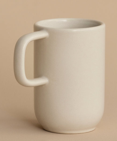 The Pacific Mug is a plain, off-white stoneware mug with a large handle and matte finish, displayed on a matching beige surface. Made in Portugal, this unadorned mug is part of the Pacific Dinnerware collection.
