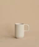The Pacific Mug, a plain white stoneware piece from Pacific Dinnerware and made in Portugal, sits slightly left of center on a beige surface with a matching background. Simple and unadorned, it offers understated elegance.