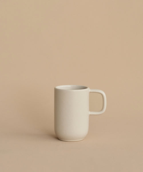 The Pacific Mug, a plain white stoneware piece from Pacific Dinnerware and made in Portugal, sits slightly left of center on a beige surface with a matching background. Simple and unadorned, it offers understated elegance.