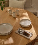 A wooden table set with stacked Pacific Dinnerware plates, a Pacific Mug (10-oz.), and a folded napkin with brown-handled cutlery. Sunlight casts shadows across the table, with a cushioned bench in the background.